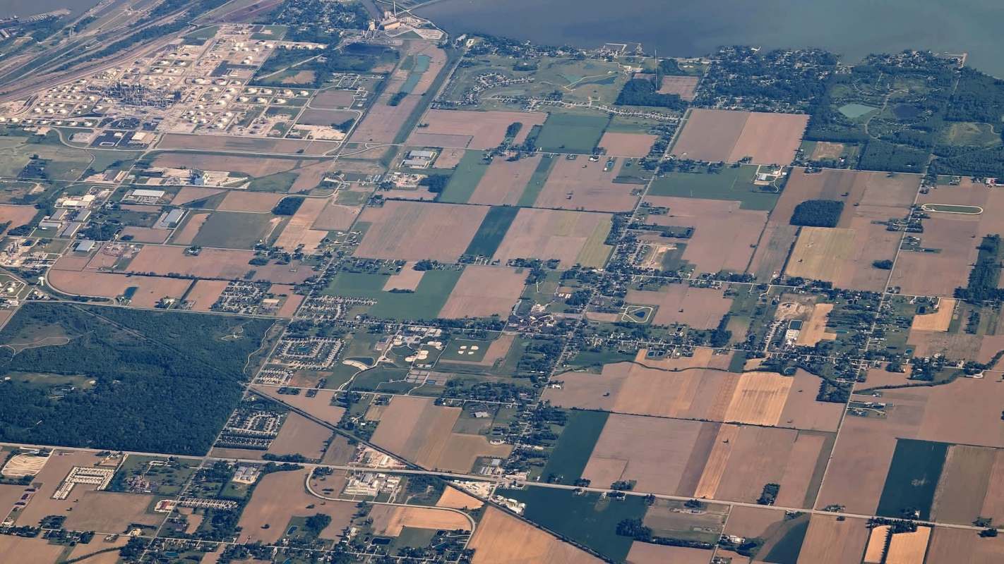 An aerial view of farmland, buildings, and roads, with a river and industrial area in the background.