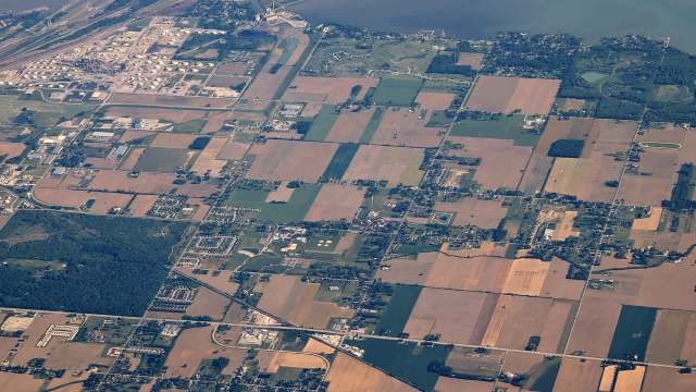 An aerial view of farmland, buildings, and roads, with a river and industrial area in the background.