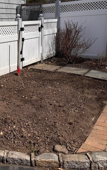 A garden area with freshly turned soil, a red-handled shovel, and stone stepping stones beside a white fence.