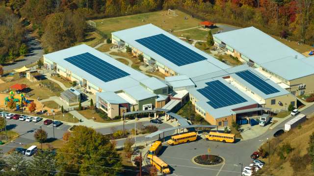Aerial view of a school with solar panels, playground, and several parked yellow school buses.