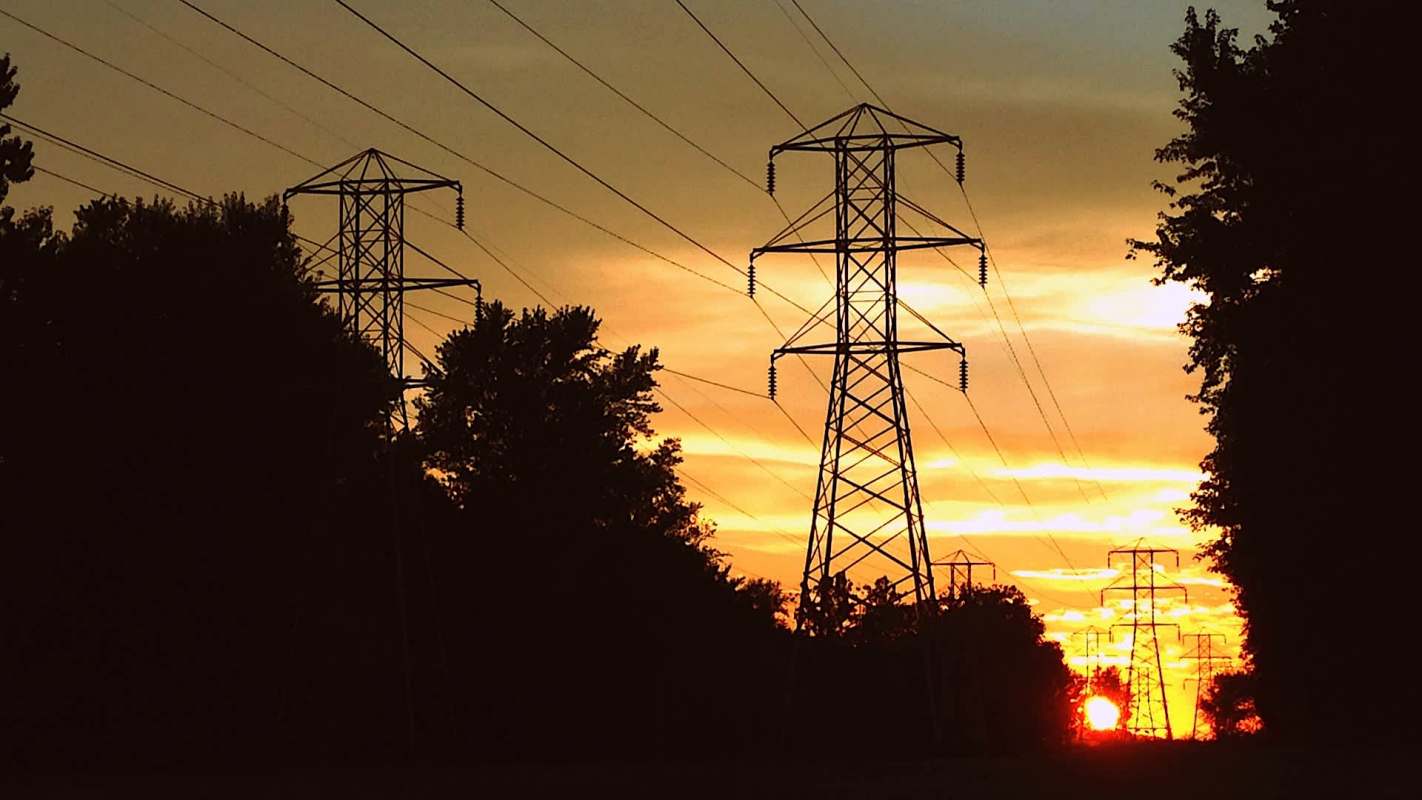 Power lines and transmission towers silhouetted against a sunset sky with vibrant orange and yellow hues.