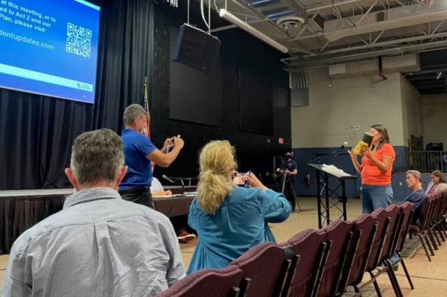 A speaker in an orange shirt addresses an audience while others use their phones to record the event.