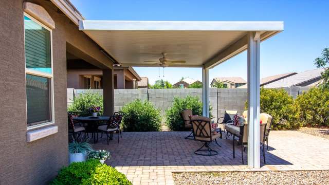 A patio area with seating under a large roof of a pergola, surrounded by greenery and a gray stone wall in a sunny setting.