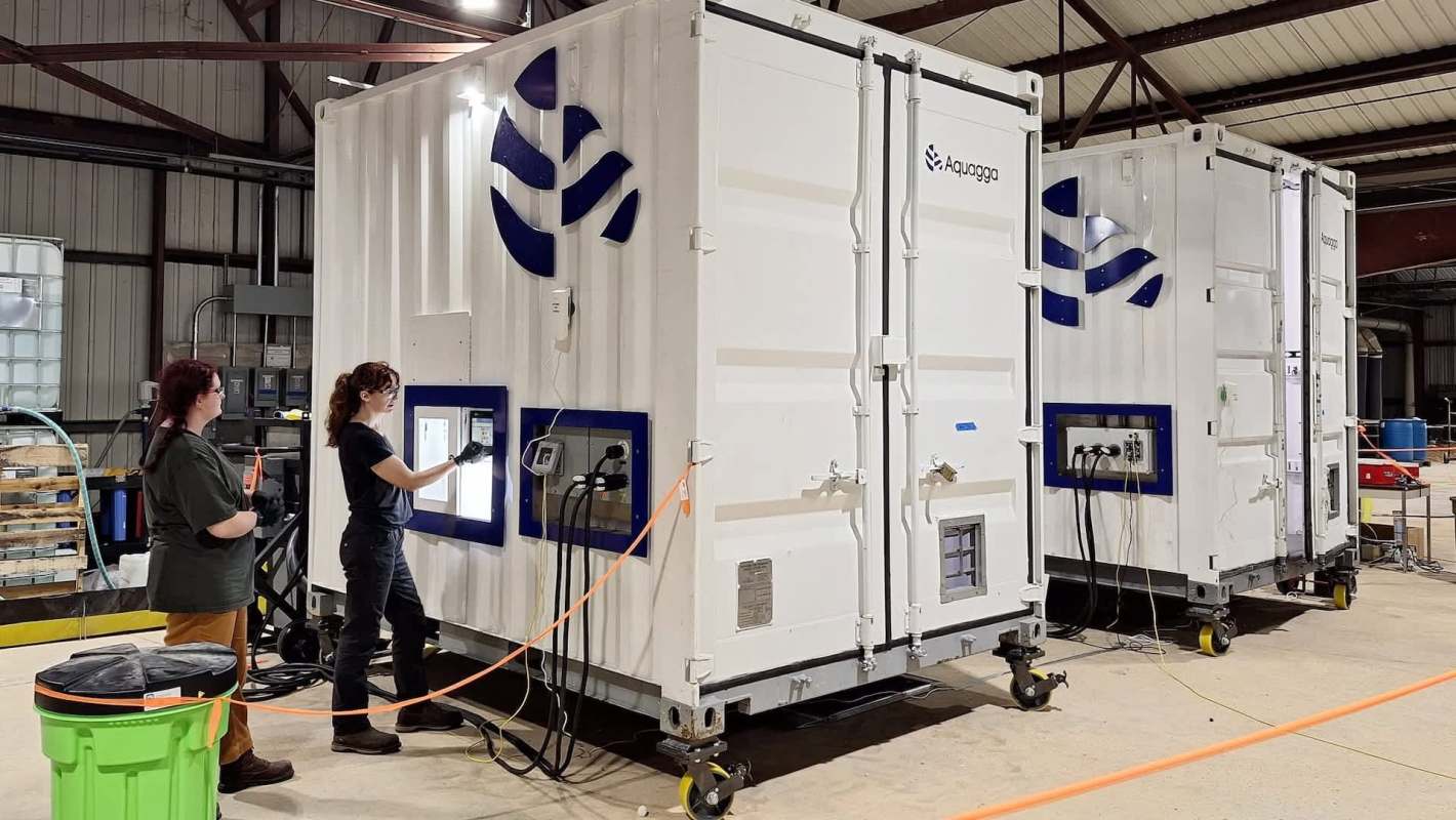 Two women work on equipment inside a warehouse, engaging with large white containers labeled with a logo.
