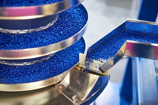 Blue plastic pellets flowing from a funnel-shaped conveyor into a collection tray.