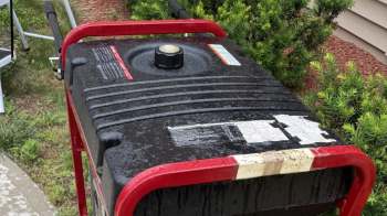 A rain-soaked portable generator with a red frame resting on a grassy area near shrubs.