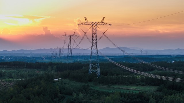 Power lines stretch across a green landscape under a colorful sunset with distant mountains.