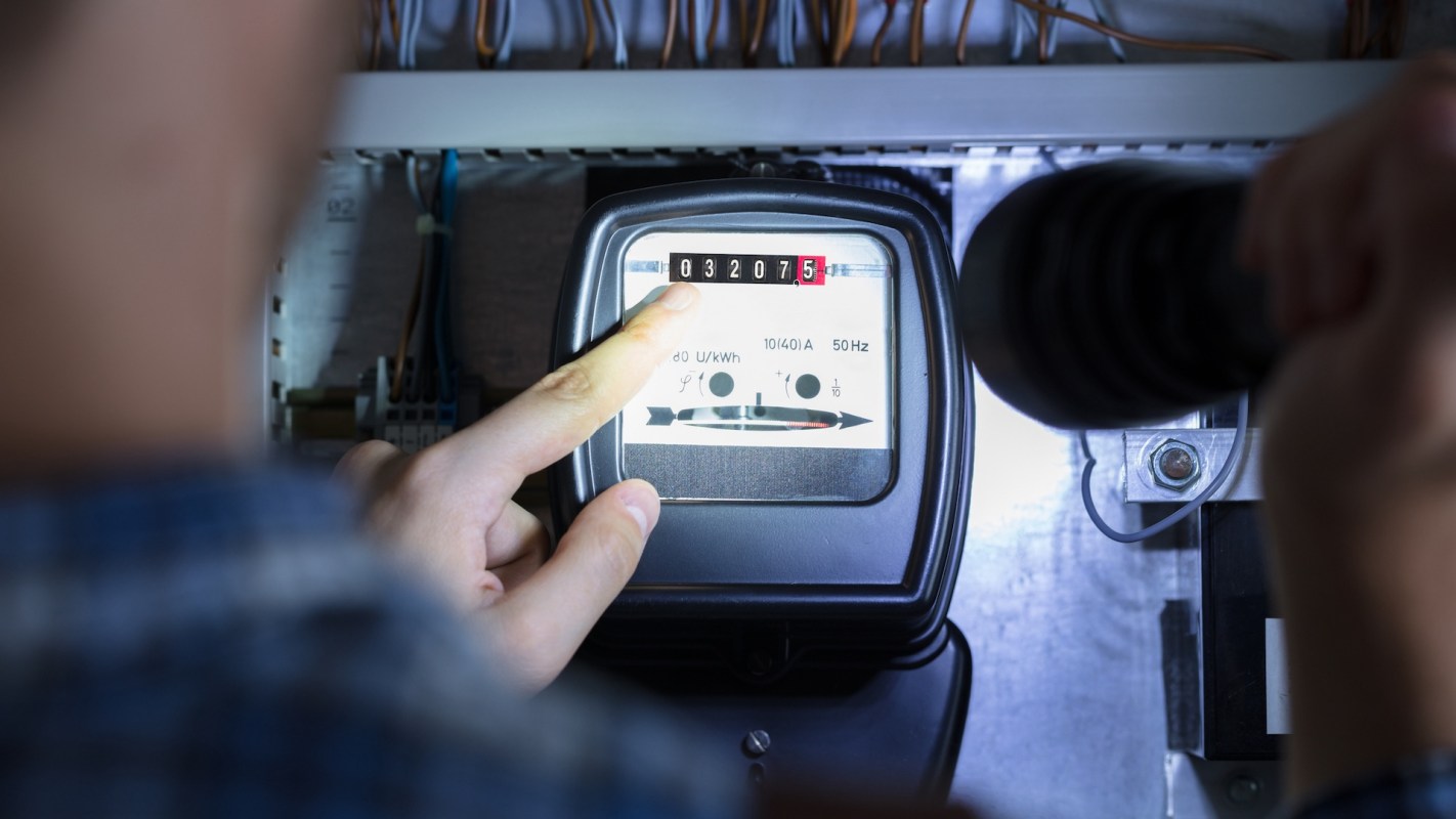 A person checks an electric meter display while using a flashlight for illumination.