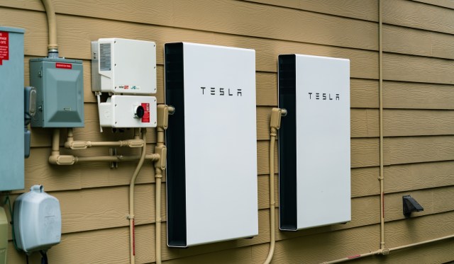 Two Tesla home battery units mounted on an exterior wall, alongside electrical components and piping.