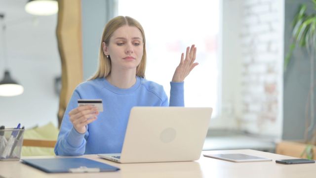 A frustrated woman holding a credit card and using a laptop.