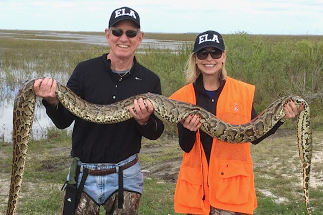 A man and woman smiling while holding a large python outdoors, with wetlands visible in the background.
