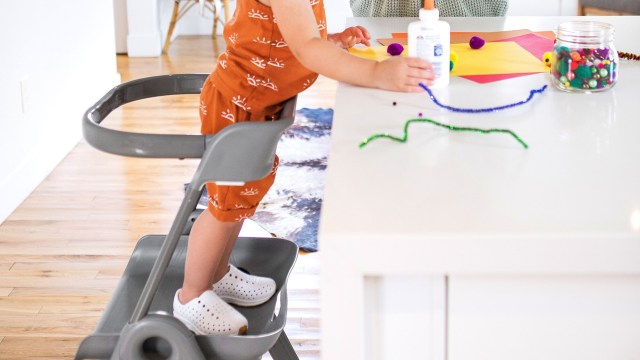 A child stands on a step stool at a table with art supplies, including glue, paper, and colorful craft materials.