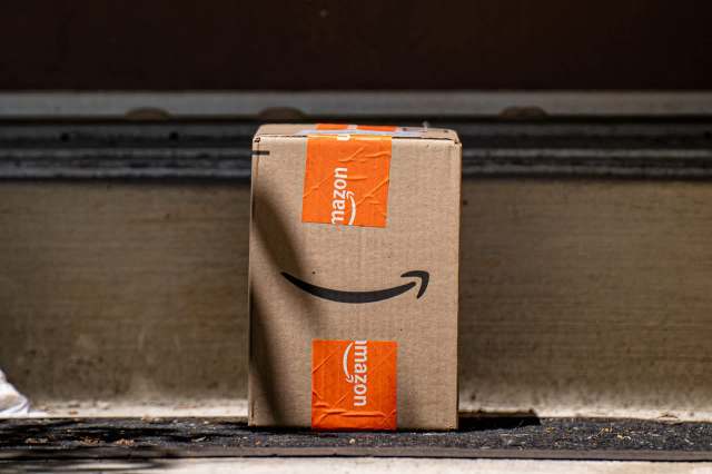 A cardboard Amazon delivery box sits on a doorstep with an orange and black logo visible.
