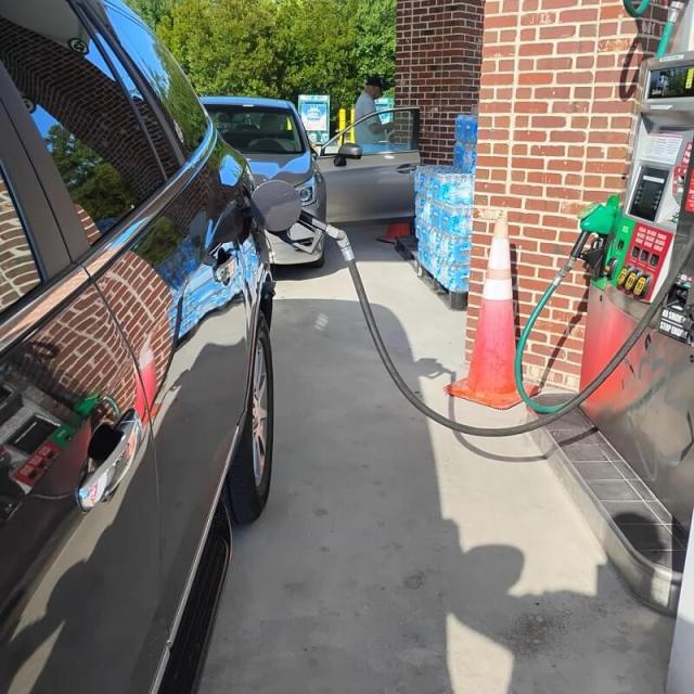 A car at a gas station with a pump nozzle inserted, alongside stacked water bottles and a person at the entrance.