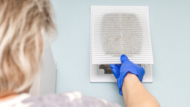 A person wearing blue gloves removes a dirty air vent cover from a wall in a light blue room.