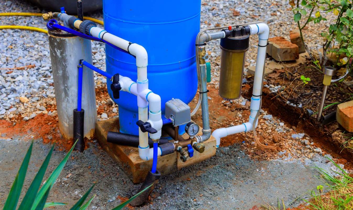 A blue water tank with connected pipes, valves, and a filtration system on a gravel surface.