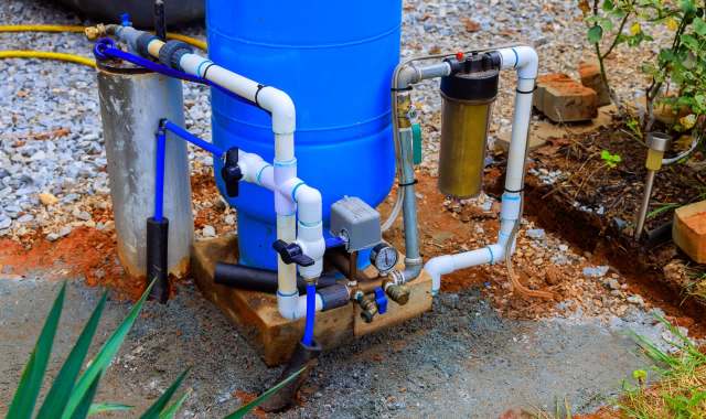 A blue water tank with connected pipes, valves, and a filtration system on a gravel surface.