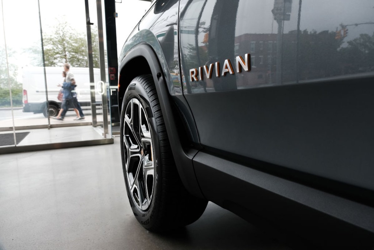 A close-up view of a Rivian vehicle's wheel and logo, with people walking outside a glass storefront in the background.