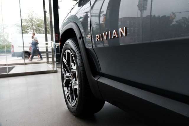 A close-up view of a Rivian vehicle's wheel and logo, with people walking outside a glass storefront in the background.