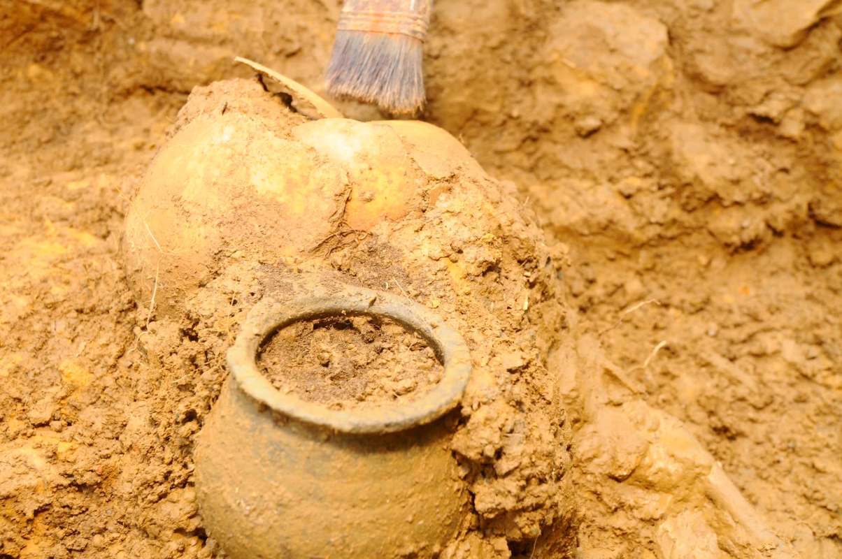 A small, partially buried pottery vessel and a larger object being gently brushed in a dirt excavation site.