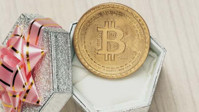 A golden bitcoin sits inside a silver gift box adorned with a pink ribbon.