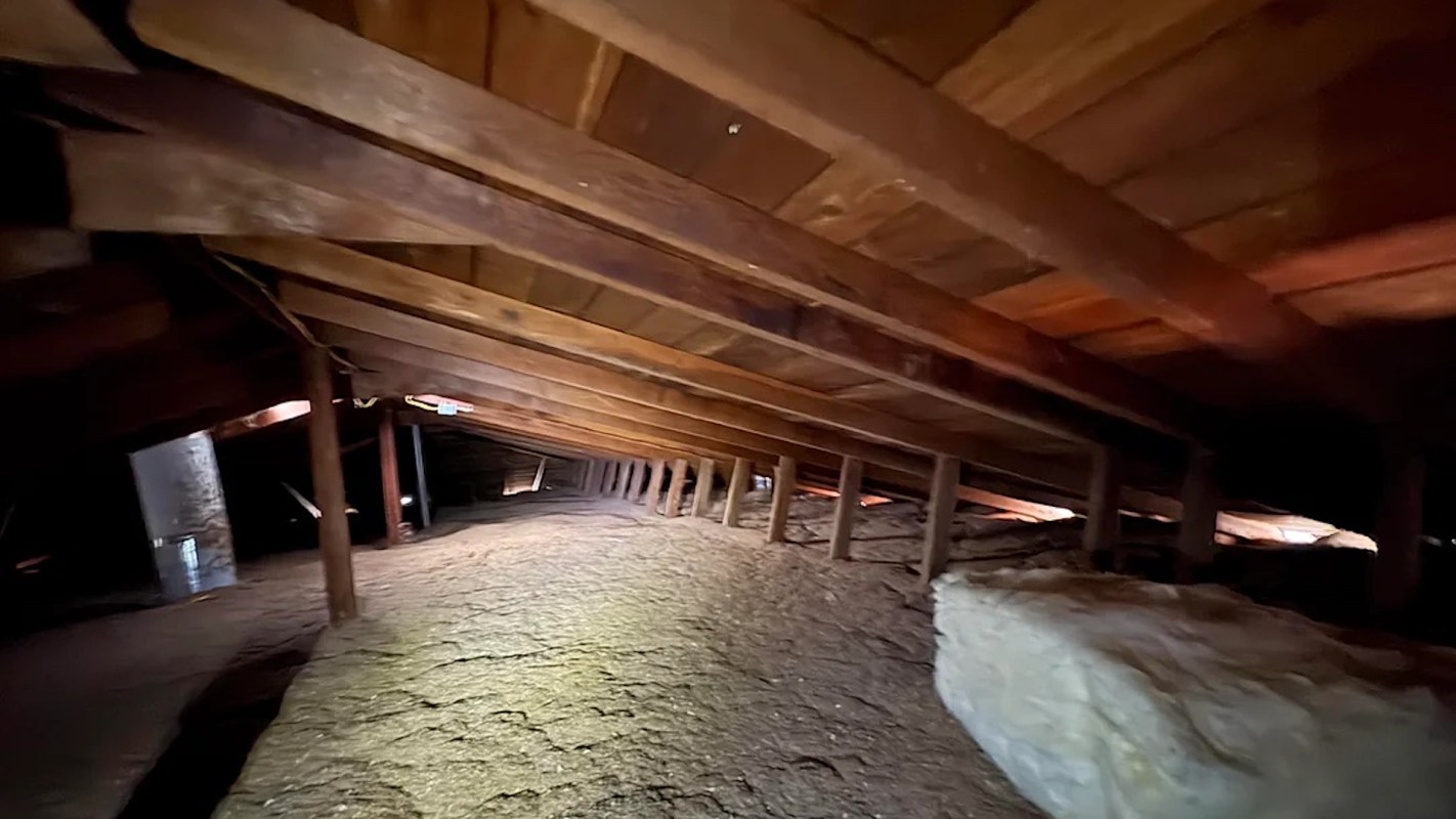 An interior view of a dark attic with wooden beams and insulation on the floor.
