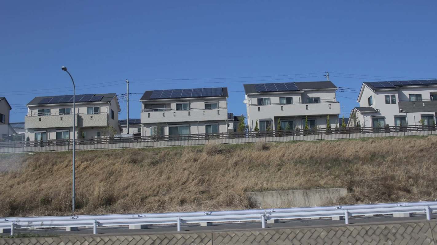 A row of modern houses with solar panels, set against a clear blue sky and grassy hillside.