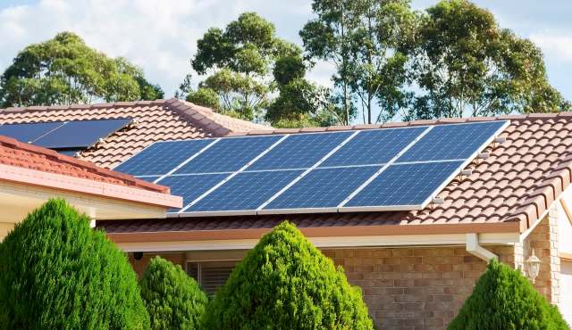 Solar panels installed on the roof of a house surrounded by green shrubs and trees.