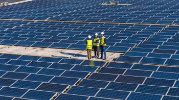Three engineers in safety vests discuss plans on a rooftop covered with solar panels.