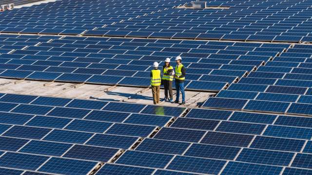 Three engineers in safety vests discuss plans on a rooftop covered with solar panels.