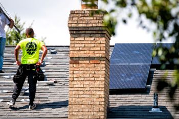 Two workers installing solar panels on a sloped roof near a brick chimney.