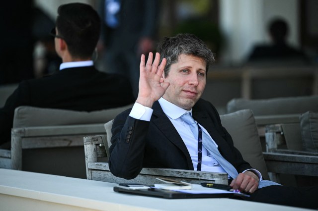 OpenAI CEO Sam Altman waves while seated on a bench with papers on a table.