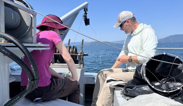 Two people on a boat engage in preparation activities on a sunny day with water and hills in the background.