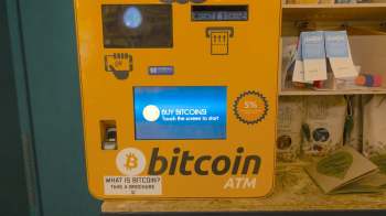 A bright yellow Bitcoin ATM with a touchscreen displaying "BUY BITCOINS! Touch the screen to start."
