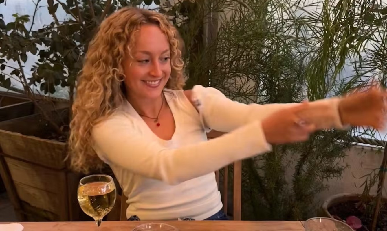 A woman smiles and tugs out a long shirt sleeve while sitting at a table with a glass of white wine.
