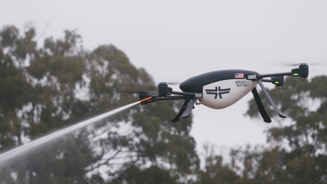 A drone equipped with a spraying mechanism flies over greenery on a cloudy day.