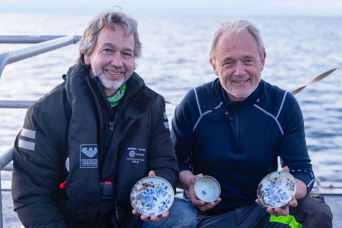 Two men holding decorative, antique plates while smiling, with a calm sea in the background.