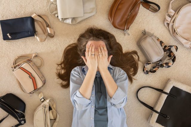 A woman lies on a carpet surrounded by various handbags, covering her face with her hands.