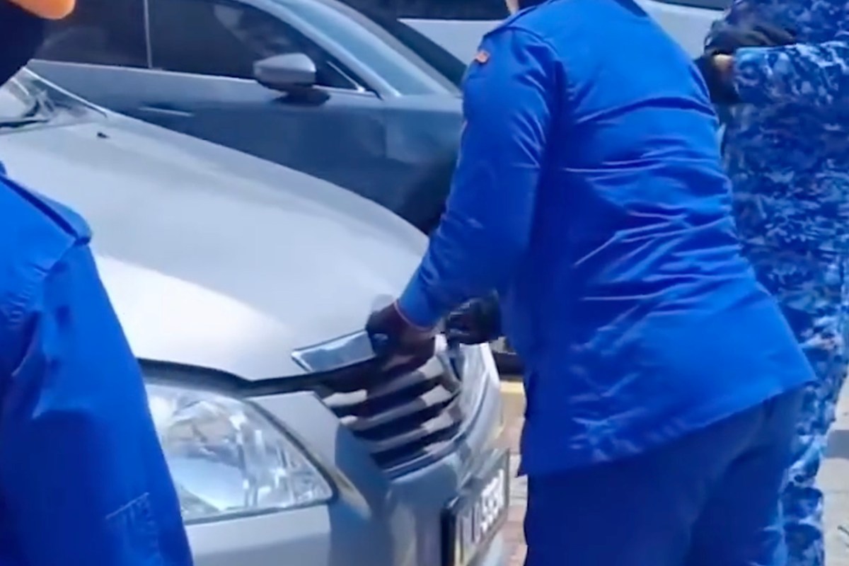 A person in a blue uniform is inspecting the front grille of a silver car.