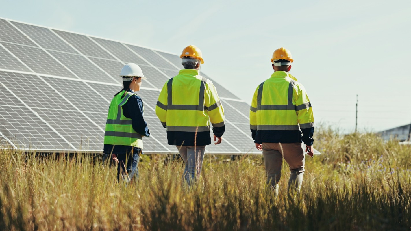 Three technicians in safety gear walk through a field with solar panels.