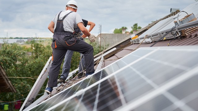 Two workers are installing solar panels on a roof under a cloudy sky.