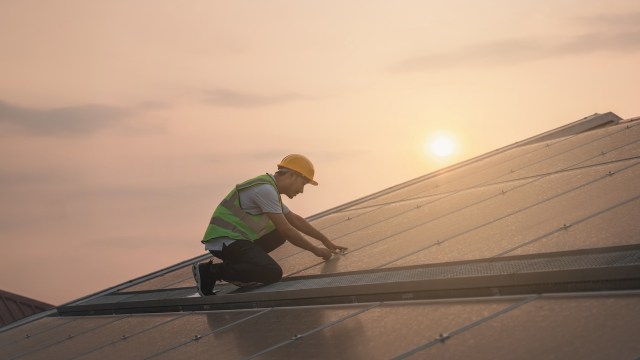A worker in a safety vest and helmet installs equipment on a solar panel rooftop at sunset.
