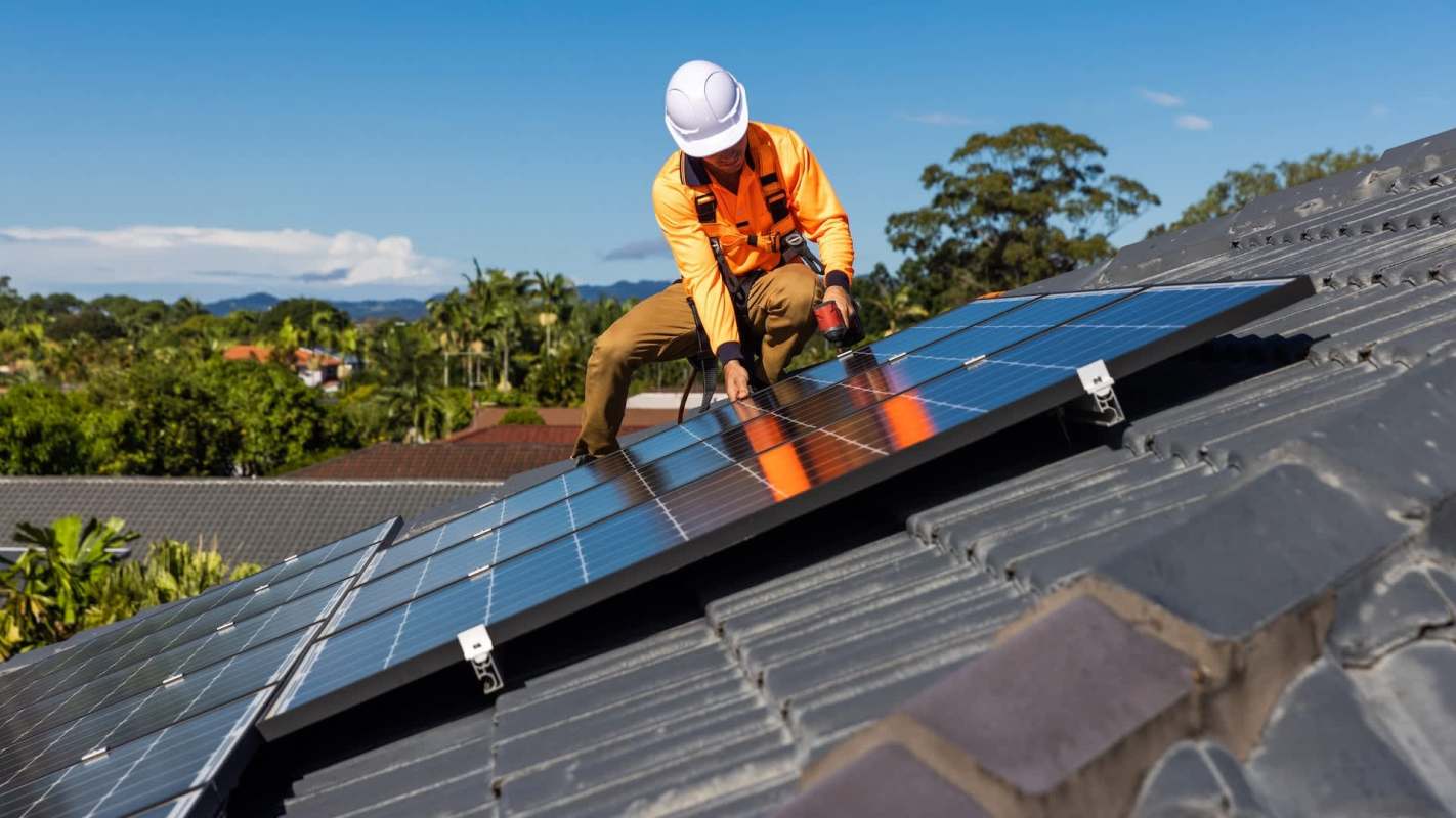 A worker in safety gear installs solar panels on a roof under a clear blue sky.