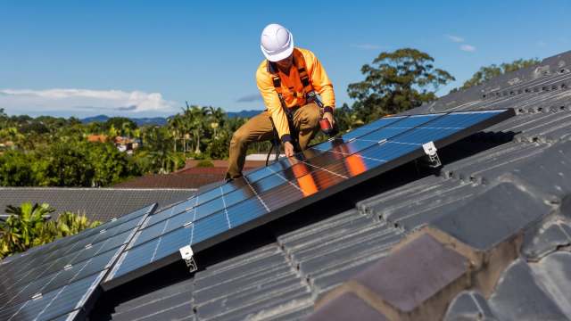 A worker in safety gear installs solar panels on a roof under a clear blue sky.