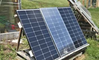 Three solar panels on a wooden stand, with the middle one with a crack, near a grassy area, with a greenhouse visible in the background.