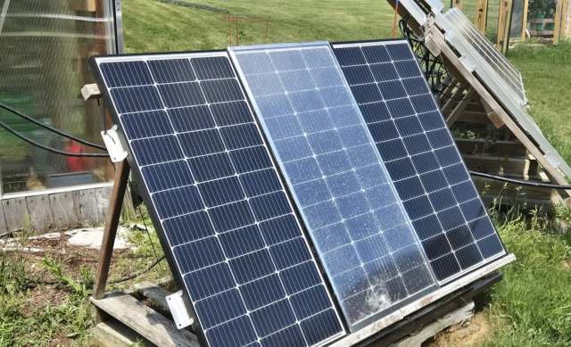 Three solar panels on a wooden stand, with the middle one with a crack, near a grassy area, with a greenhouse visible in the background.