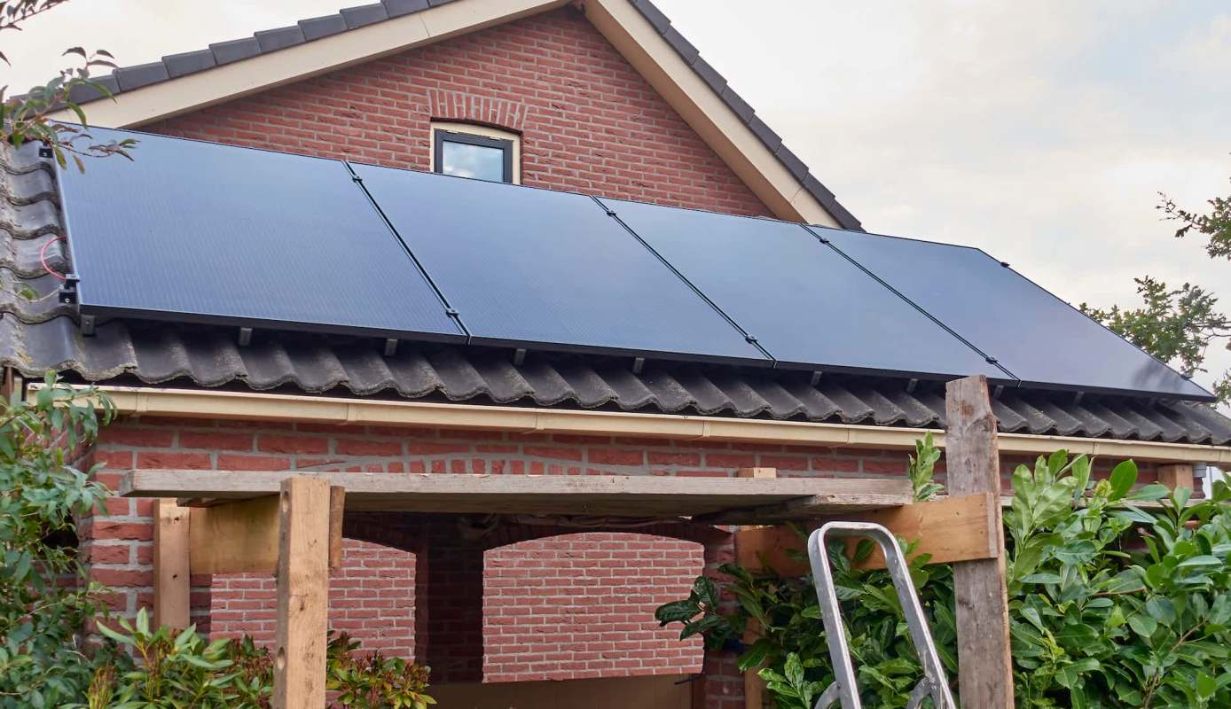 A house with solar panels installed on the roof, surrounded by greenery and a wooden structure.