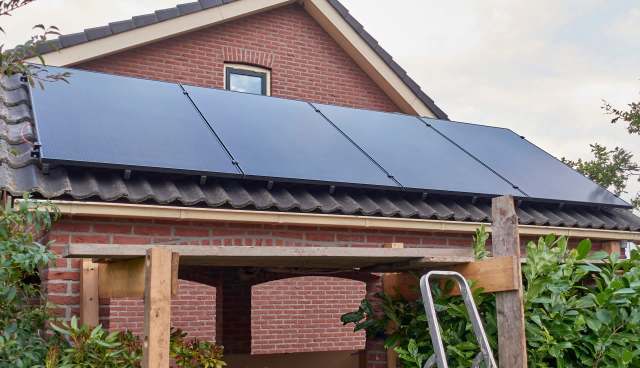 A house with solar panels installed on the roof, surrounded by greenery and a wooden structure.