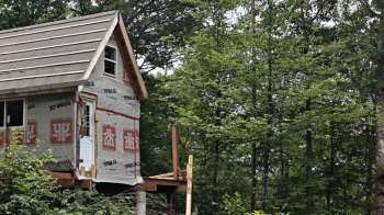 A partially constructed tiny house with a wooden deck, surrounded by lush green trees in a forested area.