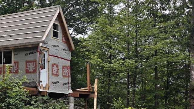 A partially constructed tiny house with a wooden deck, surrounded by lush green trees in a forested area.